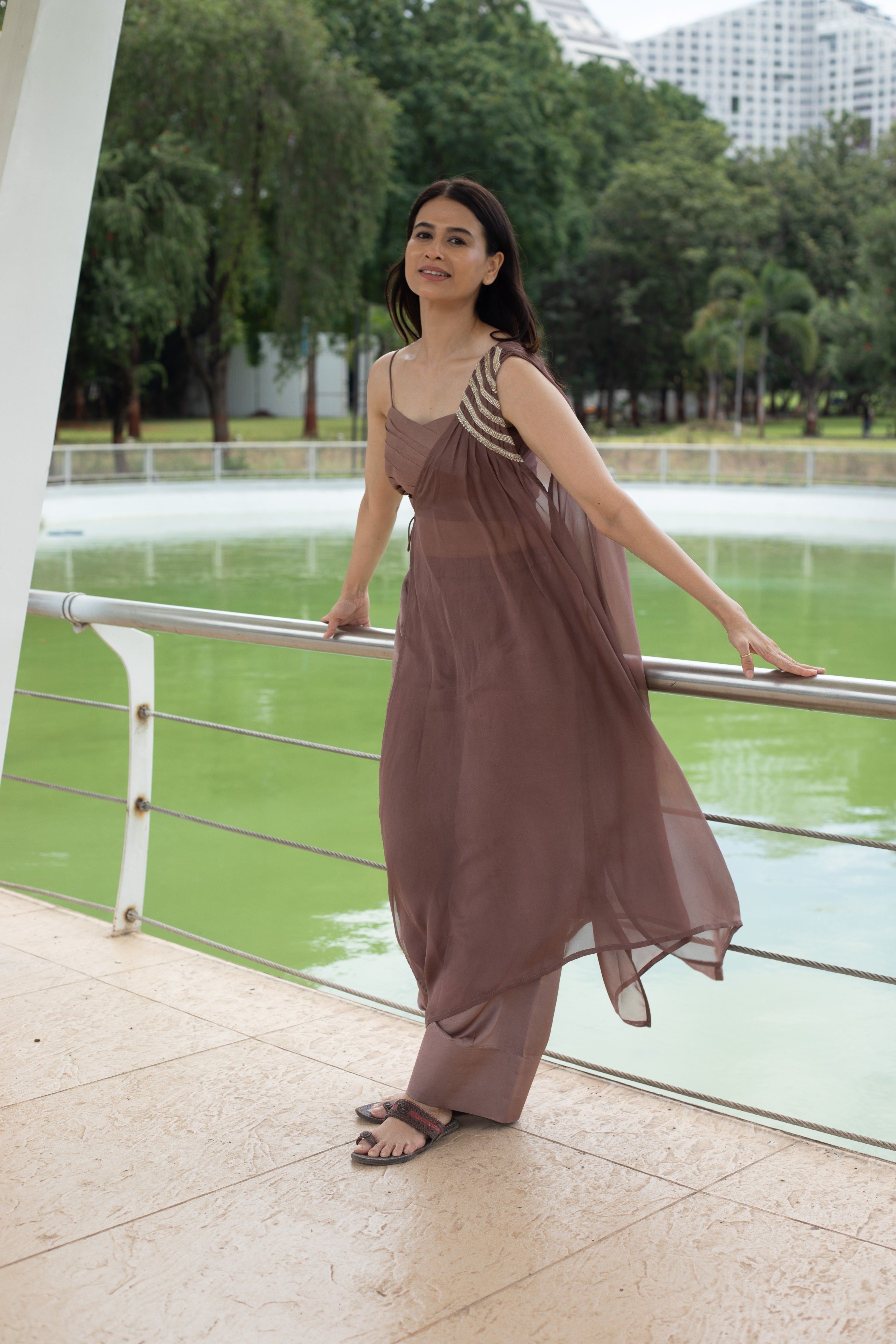 Woman in a brown dress standing by a railing with a green pond and trees in the background