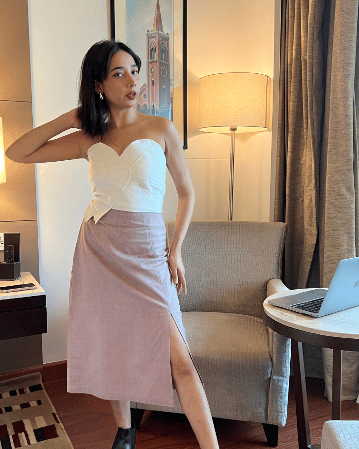 Woman in a white top and pink skirt standing in a room with a lamp and chair.
