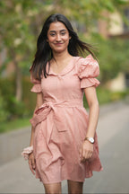 pink mini dress, peach dress, rose colored dress, Peter Pan collar dress, puff sleeve cotton dress, organic cotton clothing, sustainable vintage dress, dress with tie-belt, cottagecore aesthetic, Swaruchii fashion. A woman wearing The Rosewater Charm Dress, a short, soft-pink dress with a Peter Pan collar, puff sleeves, and a matching tie-belt, smiling in a bright, airy setting.