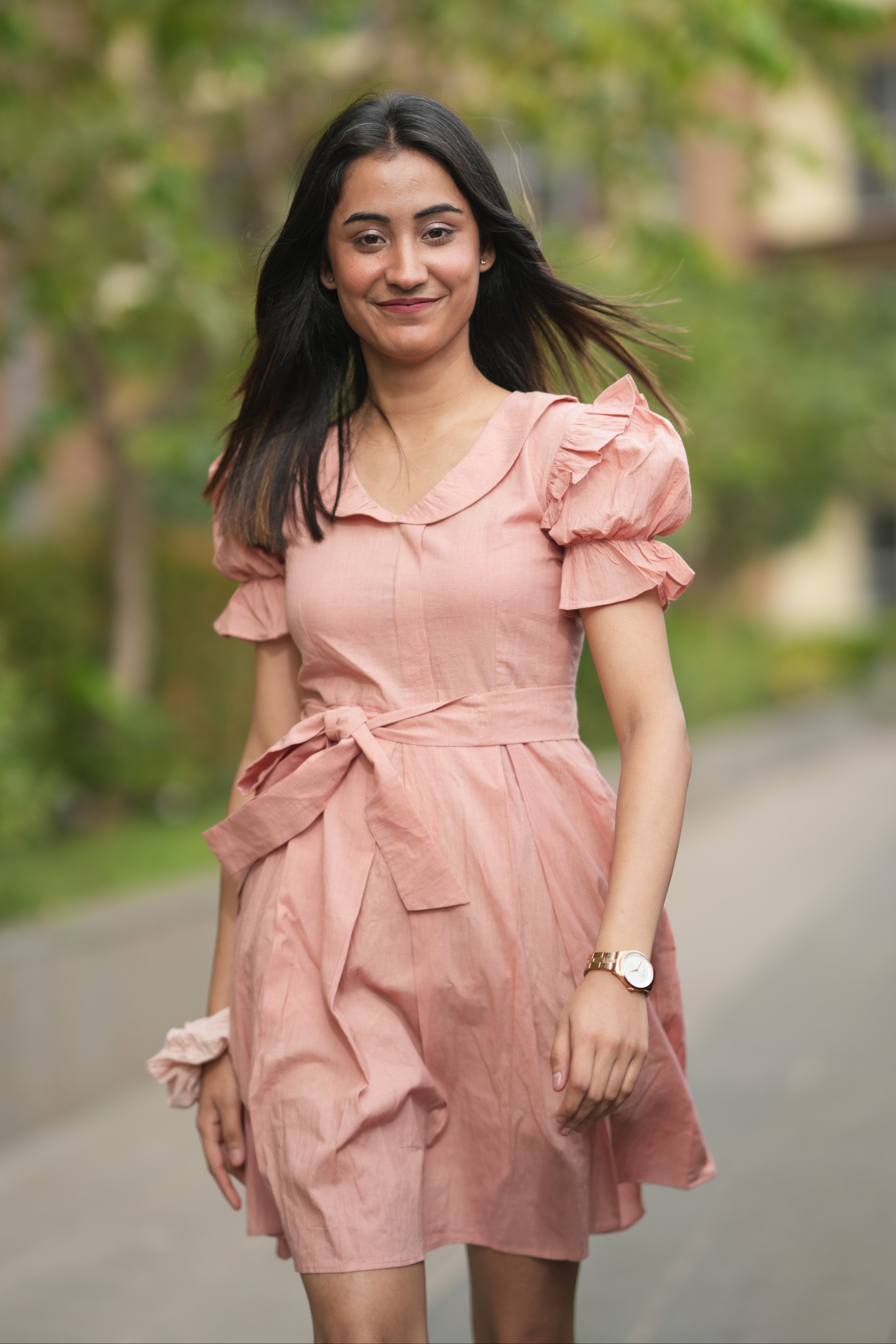 pink mini dress, peach dress, rose colored dress, Peter Pan collar dress, puff sleeve cotton dress, organic cotton clothing, sustainable vintage dress, dress with tie-belt, cottagecore aesthetic, Swaruchii fashion. A woman wearing The Rosewater Charm Dress, a short, soft-pink dress with a Peter Pan collar, puff sleeves, and a matching tie-belt, smiling in a bright, airy setting.