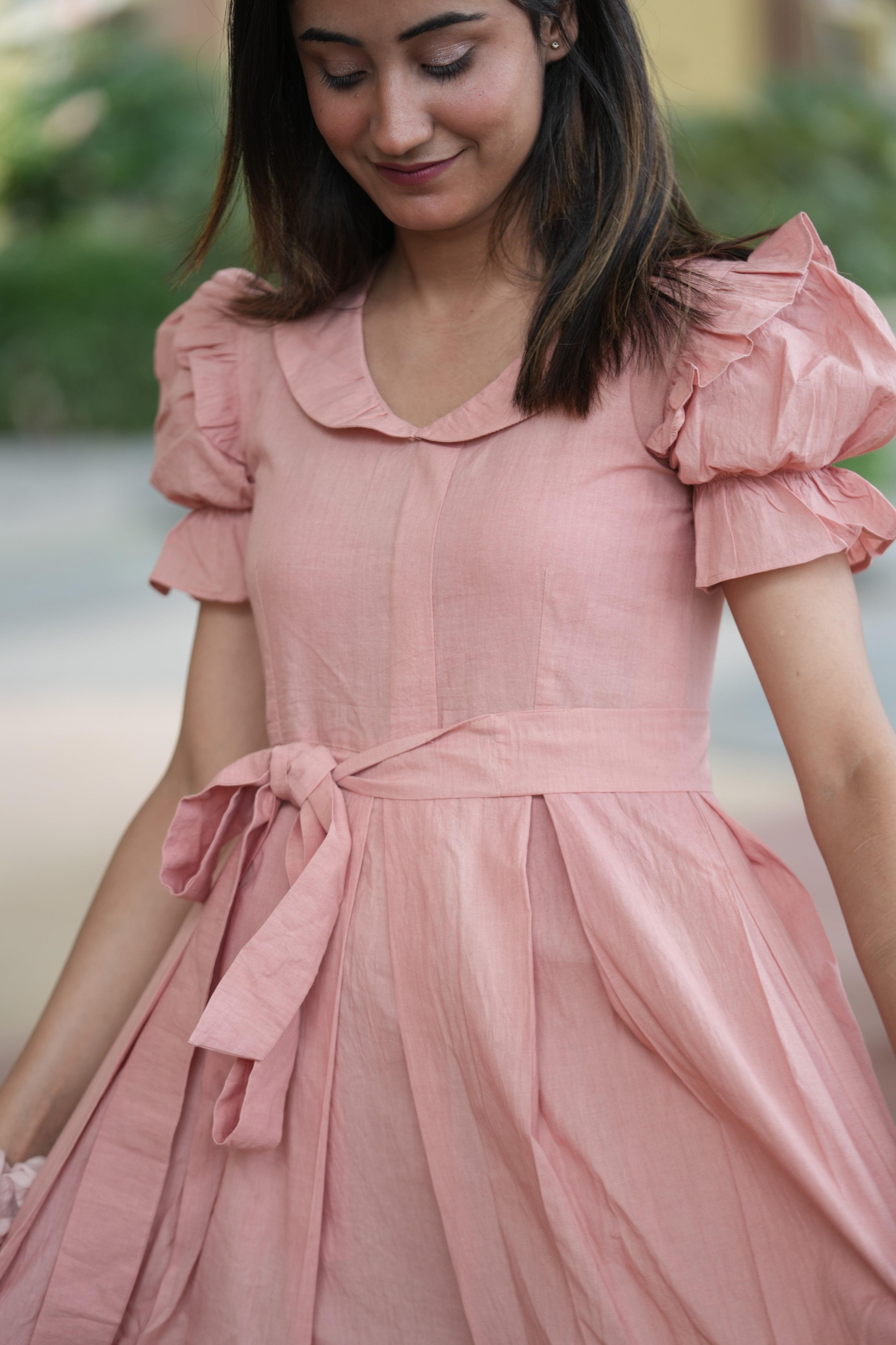 pink mini dress, peach dress, rose colored dress, Peter Pan collar dress, puff sleeve cotton dress, organic cotton clothing, sustainable vintage dress, dress with tie-belt, cottagecore aesthetic, Swaruchii fashion. A woman wearing The Rosewater Charm Dress, a short, soft-pink dress with a Peter Pan collar, puff sleeves, and a matching tie-belt, smiling in a bright, airy setting.