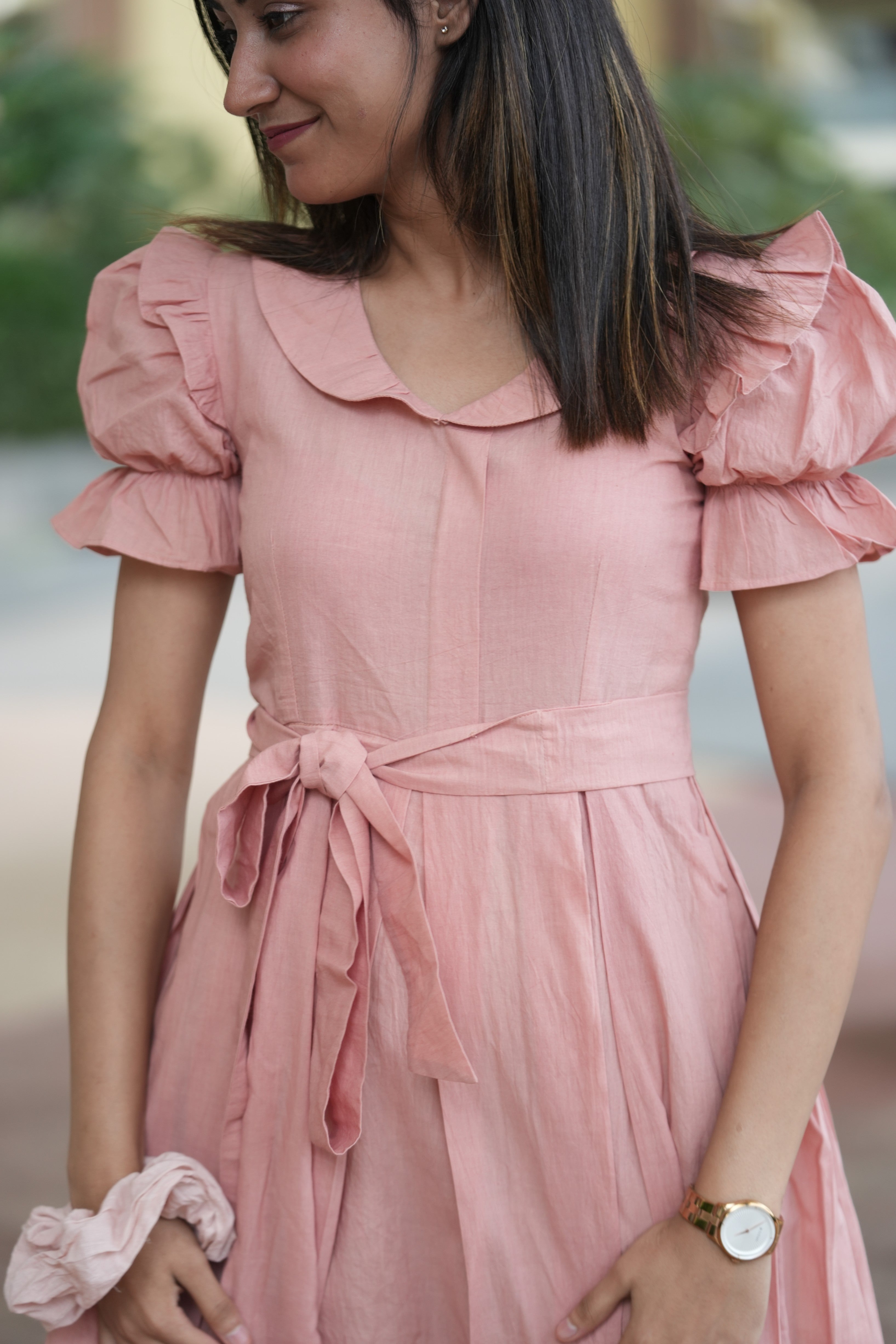 pink mini dress, peach dress, rose colored dress, Peter Pan collar dress, puff sleeve cotton dress, organic cotton clothing, sustainable vintage dress, dress with tie-belt, cottagecore aesthetic, Swaruchii fashion. A woman wearing The Rosewater Charm Dress, a short, soft-pink dress with a Peter Pan collar, puff sleeves, and a matching tie-belt, smiling in a bright, airy setting.