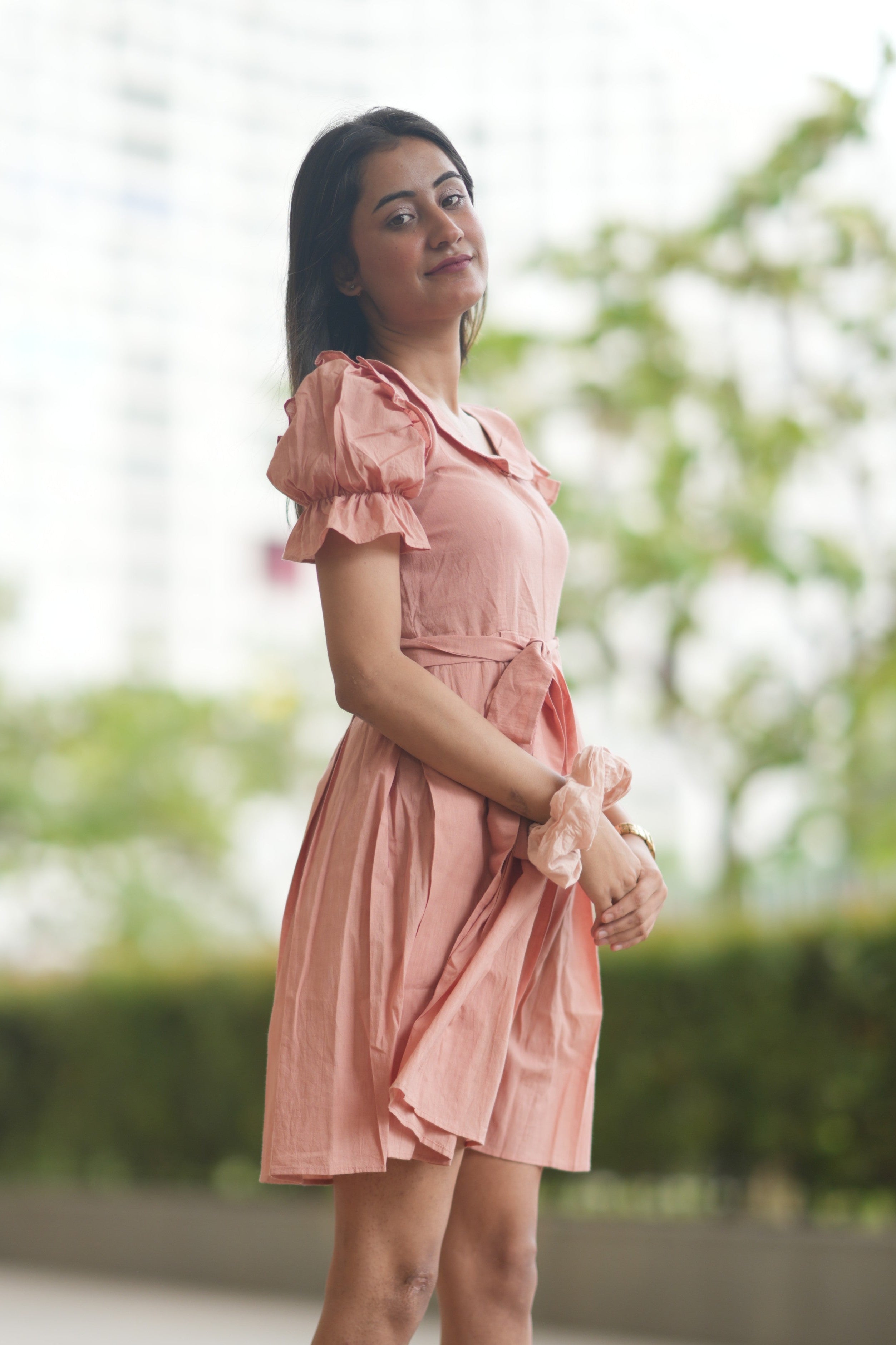 pink mini dress, peach dress, rose colored dress, Peter Pan collar dress, puff sleeve cotton dress, organic cotton clothing, sustainable vintage dress, dress with tie-belt, cottagecore aesthetic, Swaruchii fashion. A woman wearing The Rosewater Charm Dress, a short, soft-pink dress with a Peter Pan collar, puff sleeves, and a matching tie-belt, smiling in a bright, airy setting.