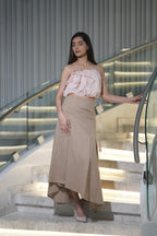 Woman in a pink top and beige skirt standing on a staircase with a modern interior background. A woman models 'The Blush' co-ord set by Swaruchii, consisting of a pink ruffled strapless crop top and a beige asymmetrical Khadi midi skirt.