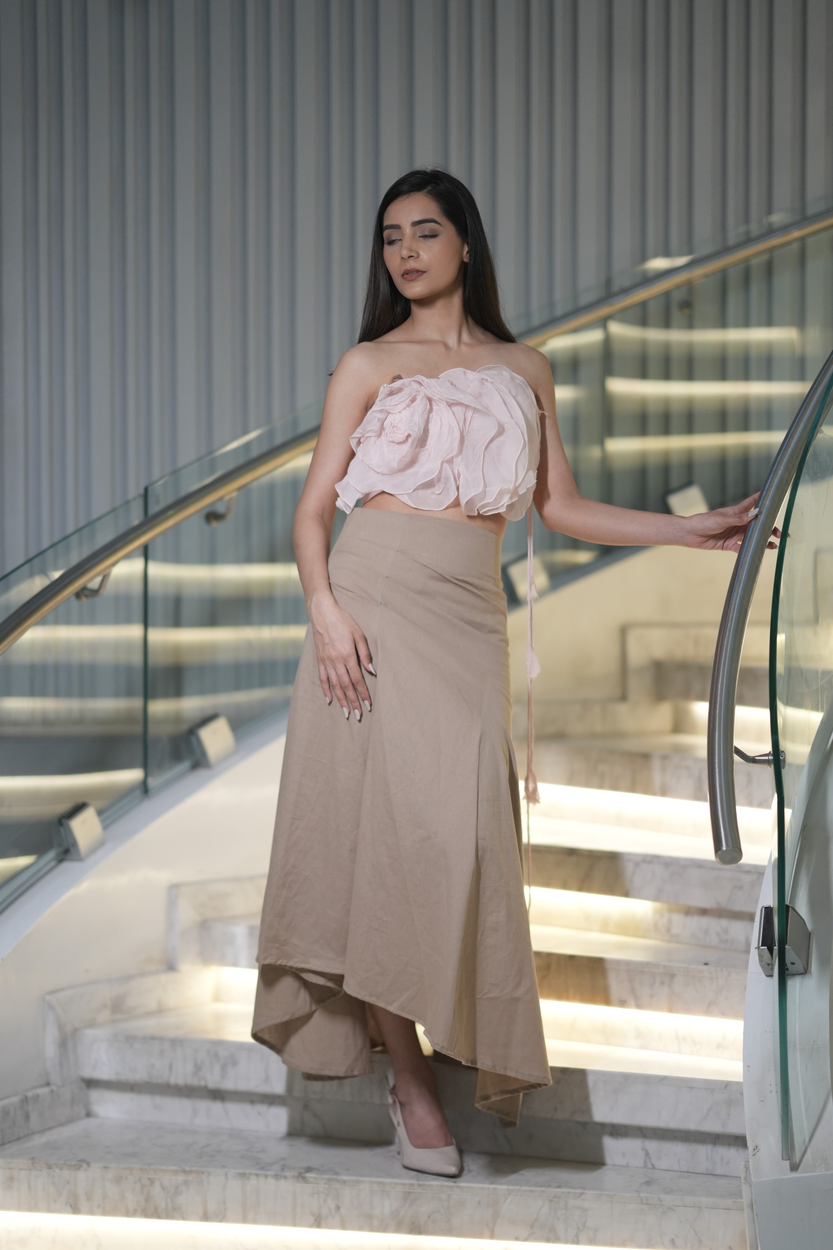 Woman in a pink top and beige skirt standing on a staircase with a modern interior background. A woman models 'The Blush' co-ord set by Swaruchii, consisting of a pink ruffled strapless crop top and a beige asymmetrical Khadi midi skirt.