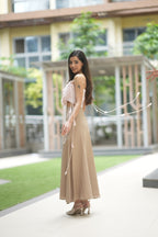 Woman in a beige dress standing outdoors with greenery and a building in the background. A woman models 'The Blush' co-ord set by Swaruchii, consisting of a pink ruffled strapless crop top and a beige asymmetrical Khadi midi skirt.