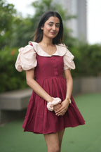 Woman wearing a maroon dress with white ruffled sleeves outdoors. red ruffle dress, organic cotton mini dress, dress with puff sleeves, waist tie dress, fit and flare dress, vintage inspired clothing, sustainable mini dress, dress with built-in shorts and pockets, Swaruchii Veira, ruby red dress. A woman wearing The Ruby Ruffle Dress, a short ruby-red dress with puff sleeves, a ruffled neckline, and a matching fabric tie-belt, twirling outdoors with a joyful expression.