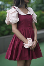 Woman wearing a maroon dress with white ruffled sleeves and holding a pink flower. red ruffle dress, organic cotton mini dress, dress with puff sleeves, waist tie dress, fit and flare dress, vintage inspired clothing, sustainable mini dress, dress with built-in shorts and pockets, Swaruchii Veira, ruby red dress.