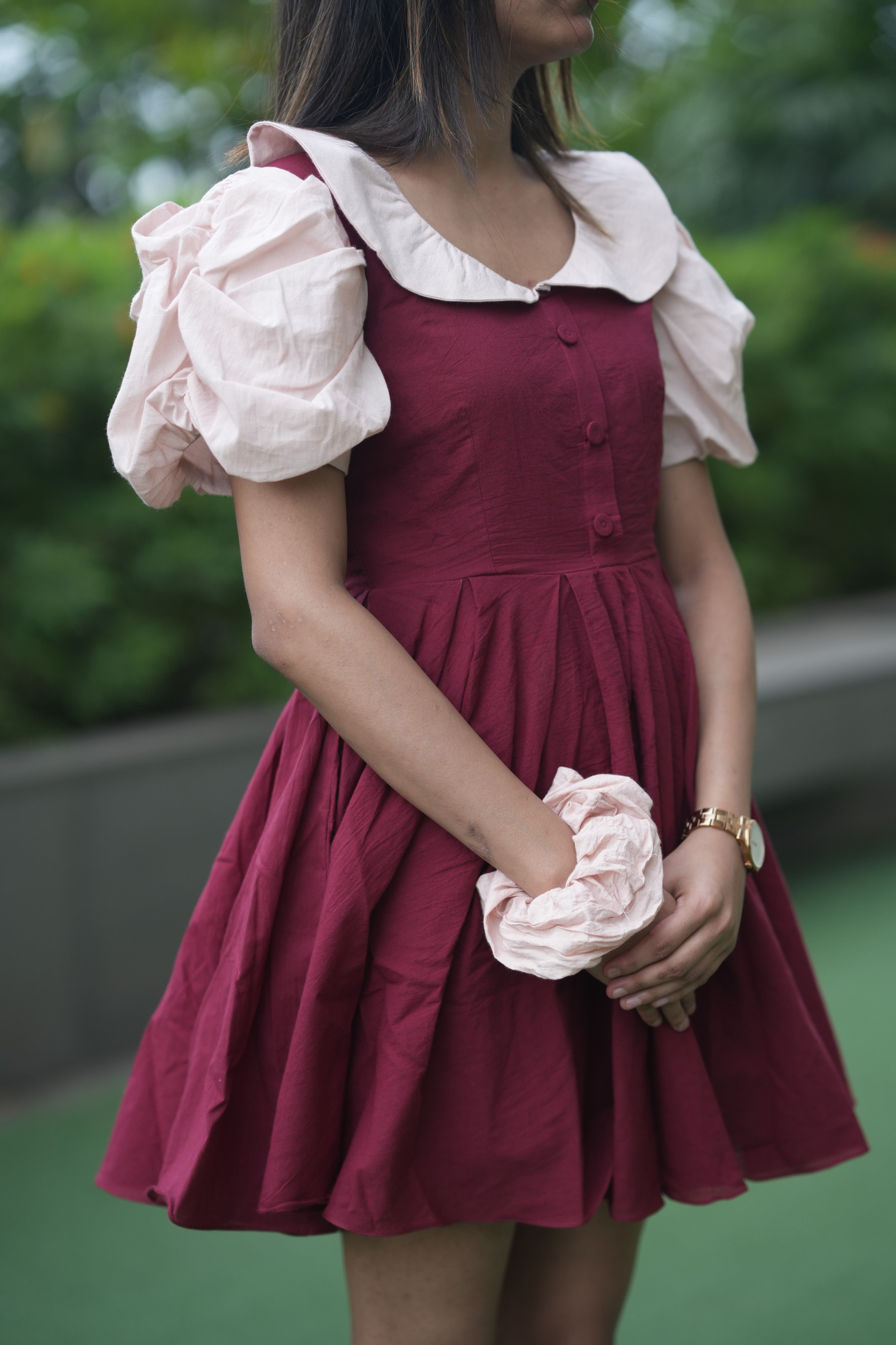 Woman wearing a maroon dress with white ruffled sleeves and holding a pink flower. red ruffle dress, organic cotton mini dress, dress with puff sleeves, waist tie dress, fit and flare dress, vintage inspired clothing, sustainable mini dress, dress with built-in shorts and pockets, Swaruchii Veira, ruby red dress.