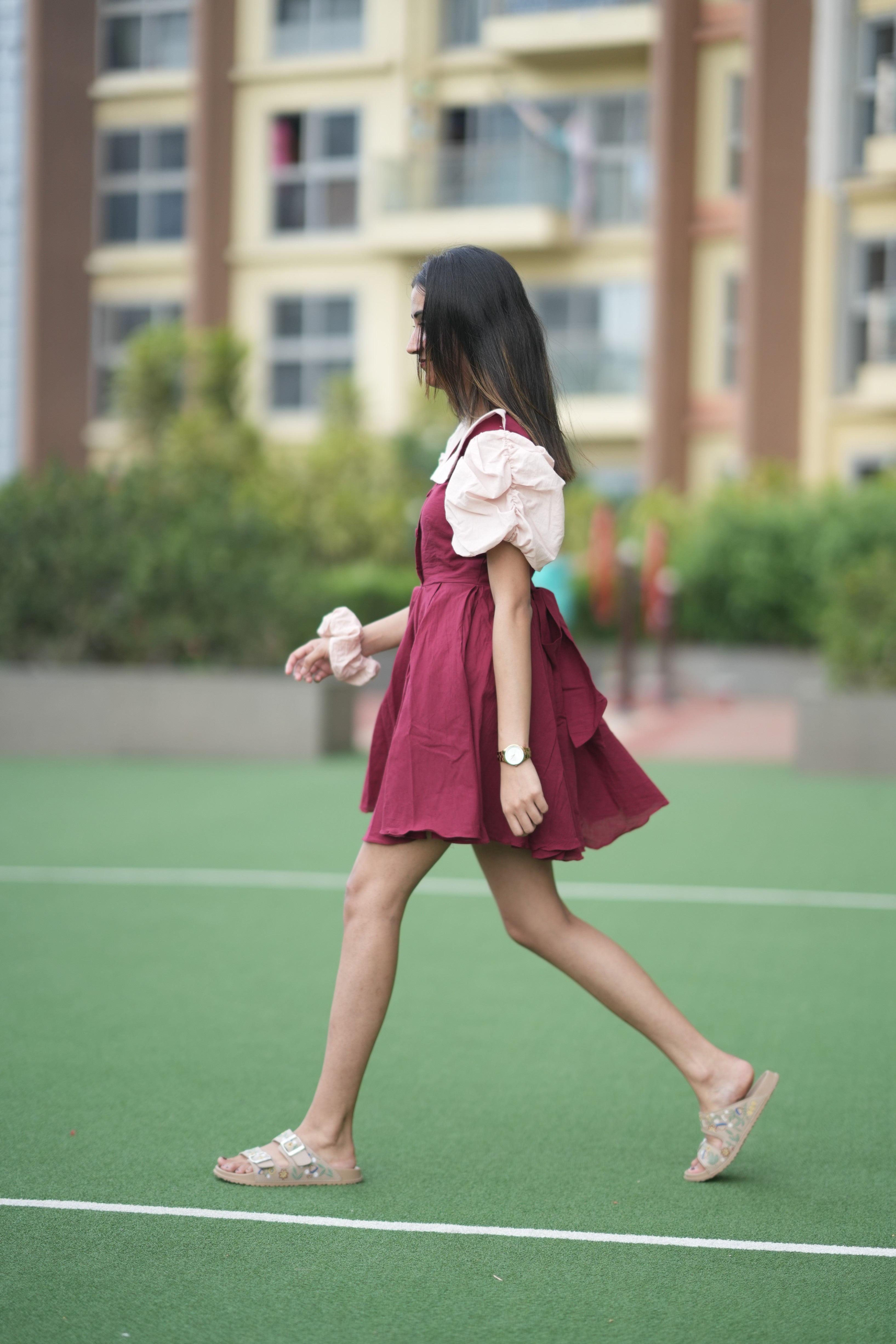 Woman in a red dress walking on a green surface with buildings in the background, red ruffle dress, organic cotton mini dress, dress with puff sleeves, waist tie dress, fit and flare dress, vintage inspired clothing, sustainable mini dress, dress with built-in shorts and pockets, Swaruchii Veira, ruby red dress.