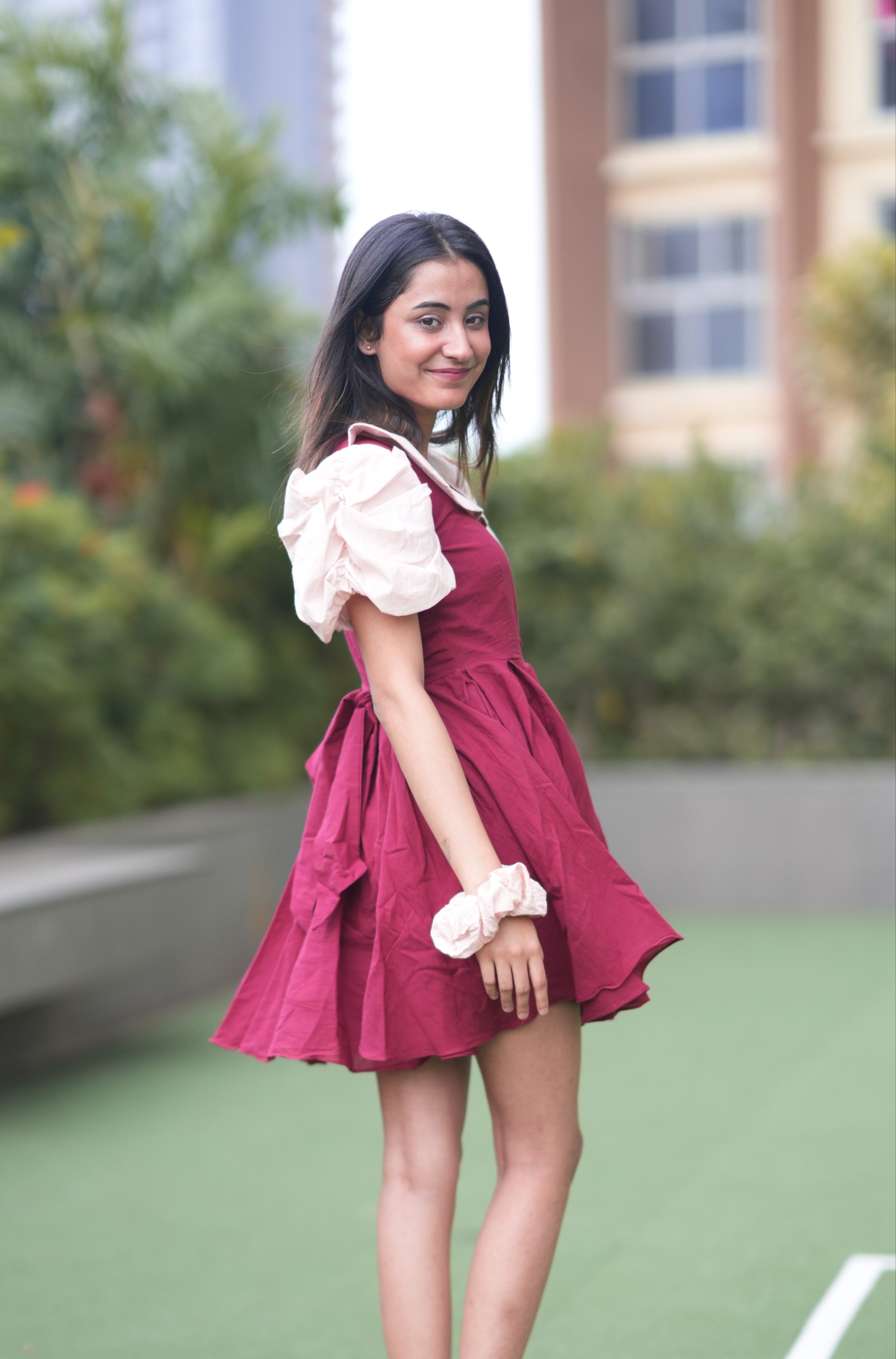 Woman in a red dress standing on a rooftop with greenery and buildings in the background. red ruffle dress, organic cotton mini dress, dress with puff sleeves, waist tie dress, fit and flare dress, vintage inspired clothing, sustainable mini dress, dress with built-in shorts and pockets, Swaruchii Veira, ruby red dress.