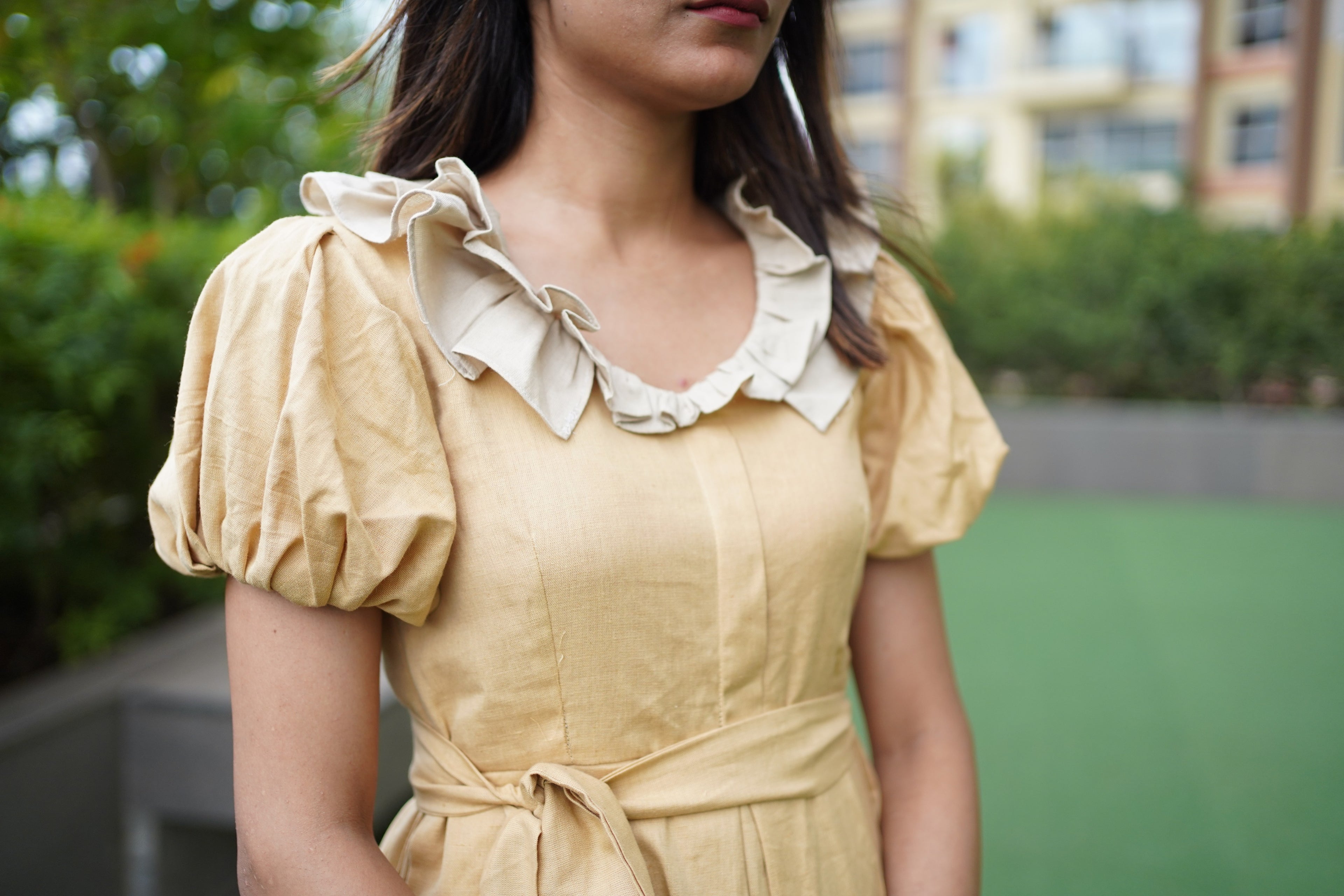 Woman wearing a yellow dress with ruffled collar in an outdoor setting. khadi dress, handwoven dress, sustainable Indian fashion, artisanal clothing, mustard yellow dress, ethical fashion, organic khadi, puff sleeve khadi dress, dress with ruffled collar, Swaruchii.