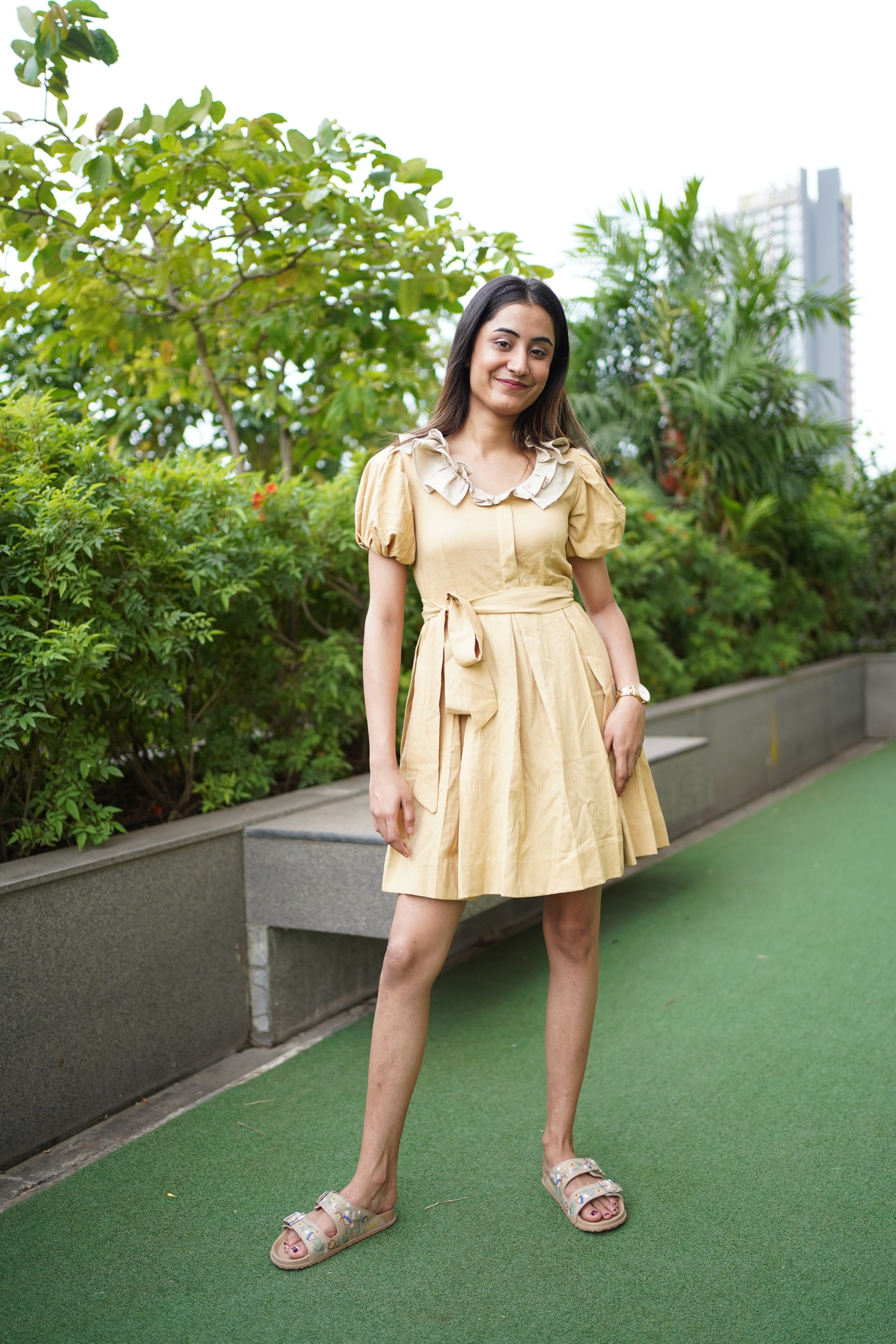 Woman in a yellow dress standing on a green rooftop with trees in the background/. khadi dress, handwoven dress, sustainable Indian fashion, artisanal clothing, mustard yellow dress, ethical fashion, organic khadi, puff sleeve khadi dress, dress with ruffled collar, Swaruchii.