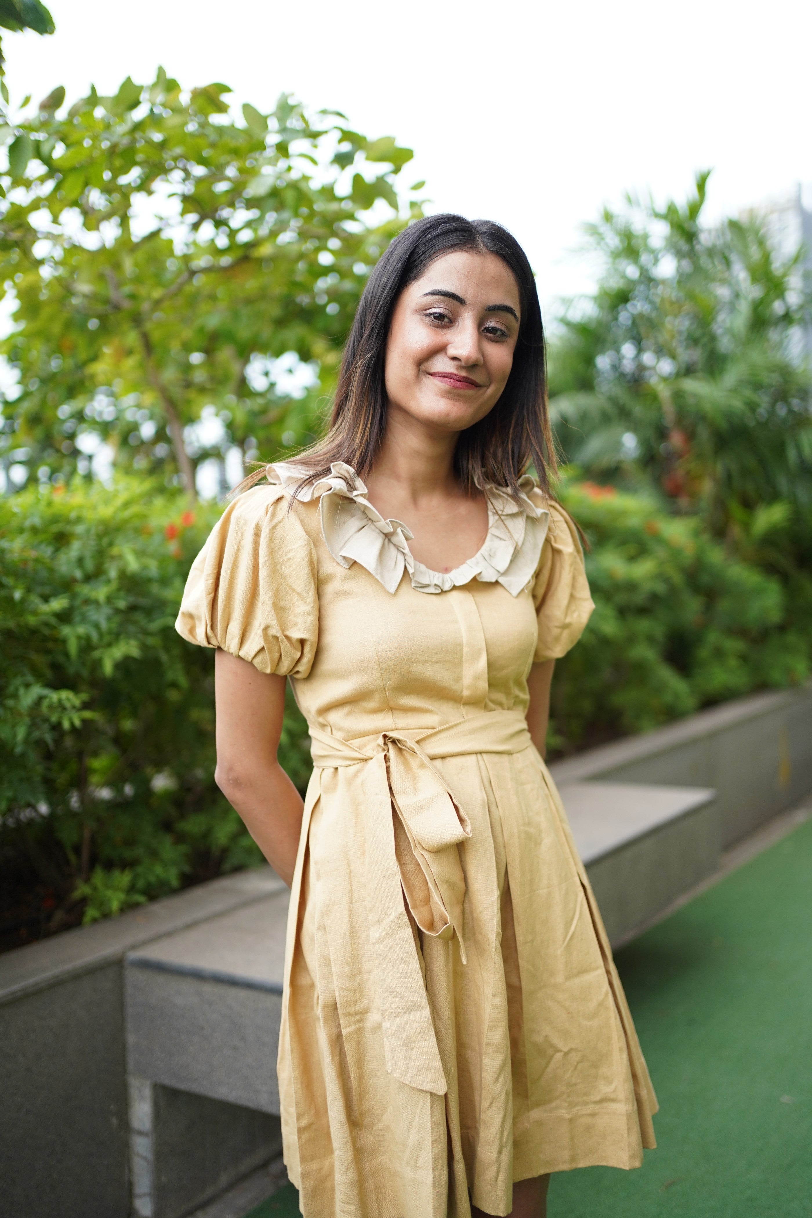 Woman in a yellow dress standing outdoors with greenery in the background. khadi dress, handwoven dress, sustainable Indian fashion, artisanal clothing, mustard yellow dress, ethical fashion, organic khadi, puff sleeve khadi dress, dress with ruffled collar, Swaruchii.