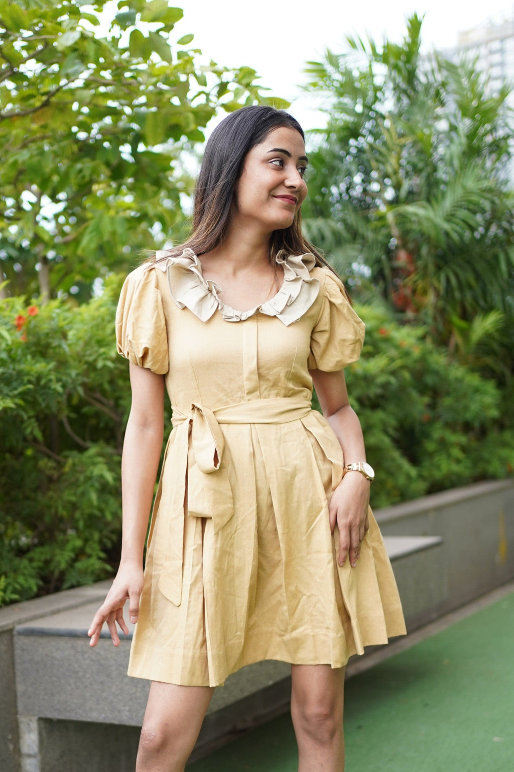 Woman in a yellow dress standing on a rooftop with greenery in the background. khadi dress, handwoven dress, sustainable Indian fashion, artisanal clothing, mustard yellow dress, ethical fashion, organic khadi, puff sleeve khadi dress, dress with ruffled collar, Swaruchii.