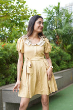 Woman in a yellow dress standing on a rooftop with greenery in the background. khadi dress, handwoven dress, sustainable Indian fashion, artisanal clothing, mustard yellow dress, ethical fashion, organic khadi, puff sleeve khadi dress, dress with ruffled collar, Swaruchii.