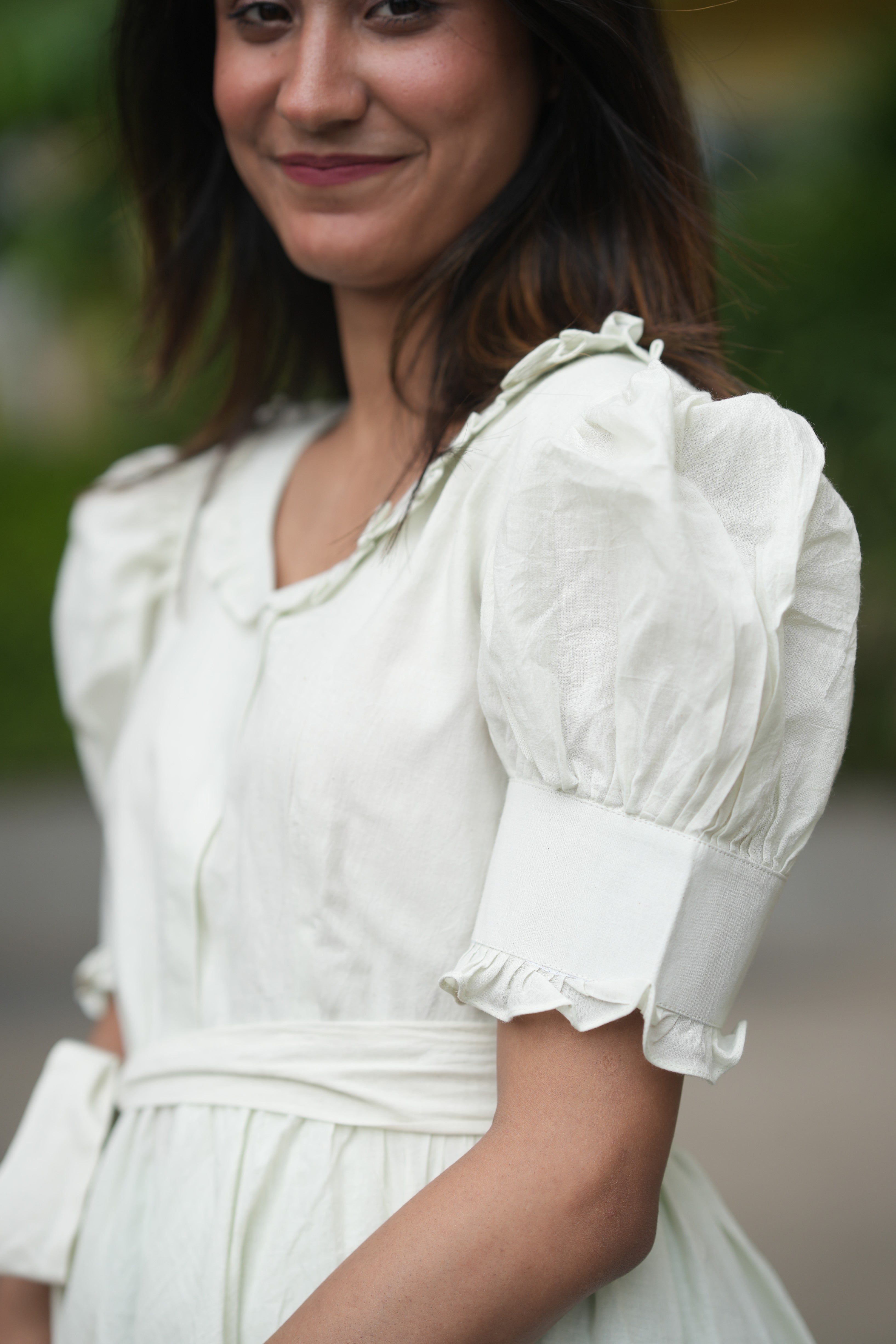 Woman wearing a white dress with puffed sleeves against a blurred outdoor background. light green dress, mint green mini dress, pastel dress, organic cotton puff sleeve dress, sustainable clothing brand, vintage style short dress, dress with pockets and shorts, eco-friendly fashion, Swaruchii Veira, meadow dress. A woman wearing The Mint Meadow Dress, a short dress in a light mint green shade with puff sleeves and a matching fabric tie-belt, standing against a lush green background.