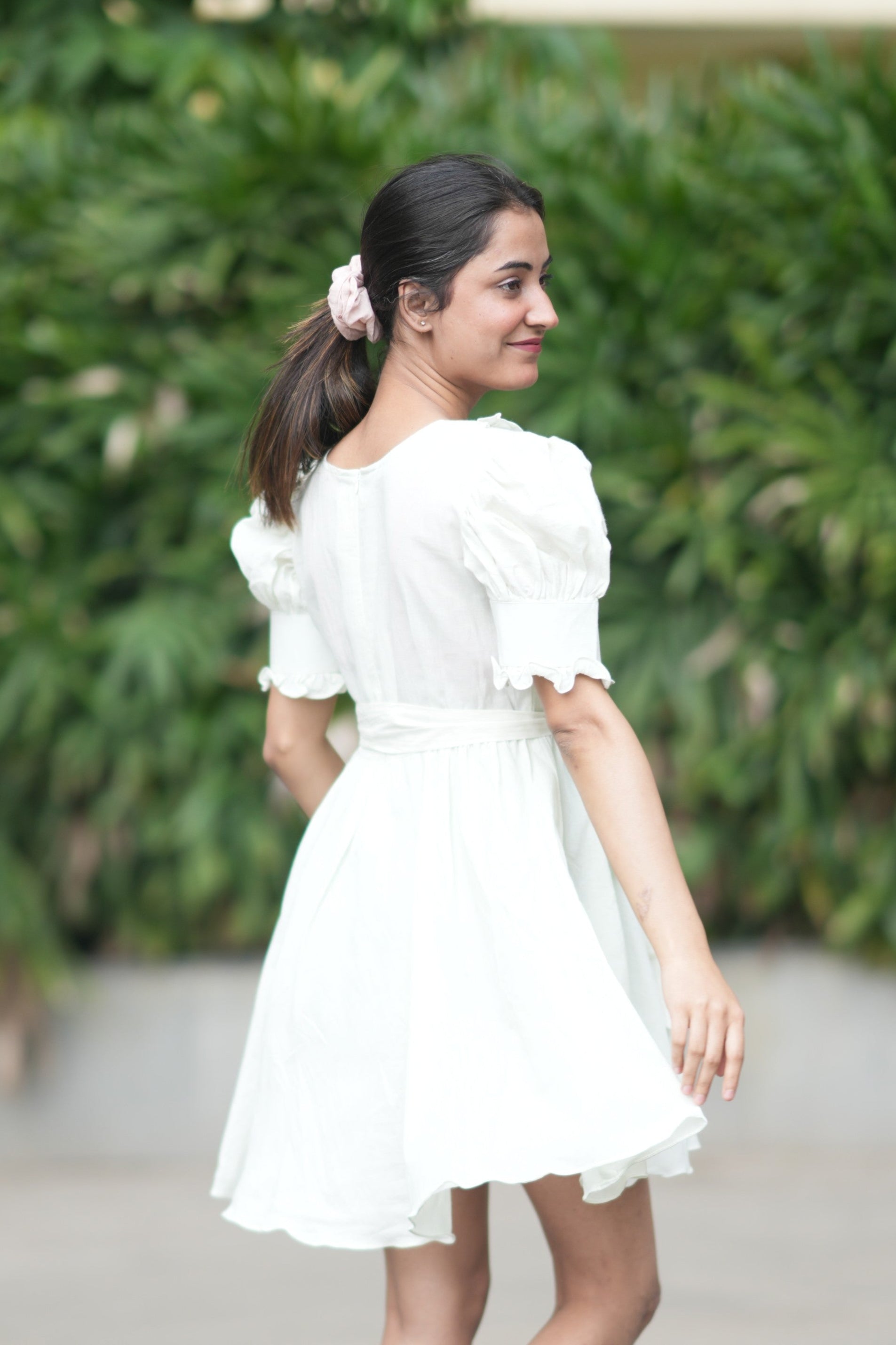 Woman in a white dress standing outdoors with greenery in the background. light green dress, mint green mini dress, pastel dress, organic cotton puff sleeve dress, sustainable clothing brand, vintage style short dress, dress with pockets and shorts, eco-friendly fashion, Swaruchii Veira, meadow dress. A woman wearing The Mint Meadow Dress, a short dress in a light mint green shade with puff sleeves and a matching fabric tie-belt, standing against a lush green background.