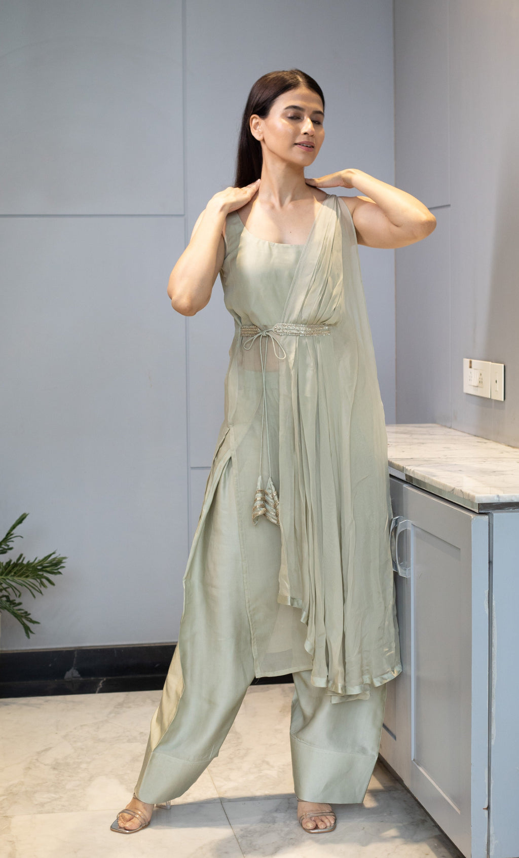 Woman in a light green dress standing in a modern indoor setting with a plant and geometric wall design.