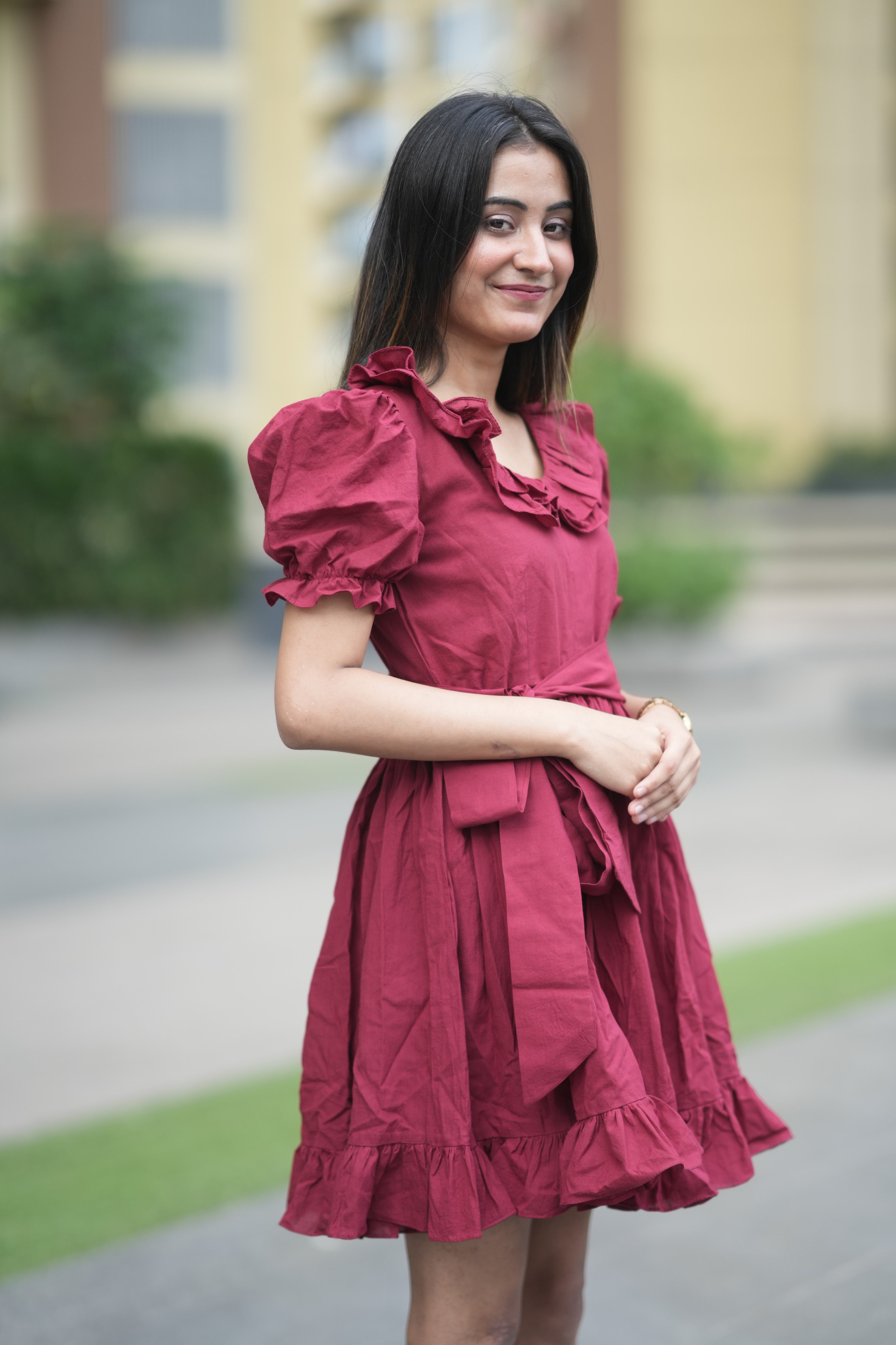 red ruffle dress, organic cotton mini dress, dress with puff sleeves, waist tie dress, fit and flare dress, vintage inspired clothing, sustainable mini dress, dress with built-in shorts and pockets, Swaruchii Veira, ruby red dress. A woman wearing The Ruby Ruffle Dress, a short ruby-red dress with puff sleeves, a ruffled neckline, and a matching fabric tie-belt, twirling outdoors with a joyful expression.