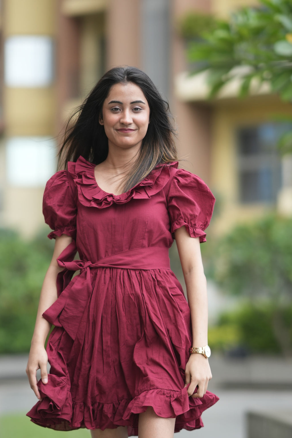 red ruffle dress, organic cotton mini dress, dress with puff sleeves, waist tie dress, fit and flare dress, vintage inspired clothing, sustainable mini dress, dress with built-in shorts and pockets, Swaruchii Veira, ruby red dress. A woman wearing The Ruby Ruffle Dress, a short ruby-red dress with puff sleeves, a ruffled neckline, and a matching fabric tie-belt, twirling outdoors with a joyful expression.