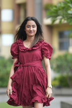 red ruffle dress, organic cotton mini dress, dress with puff sleeves, waist tie dress, fit and flare dress, vintage inspired clothing, sustainable mini dress, dress with built-in shorts and pockets, Swaruchii Veira, ruby red dress. A woman wearing The Ruby Ruffle Dress, a short ruby-red dress with puff sleeves, a ruffled neckline, and a matching fabric tie-belt, twirling outdoors with a joyful expression.