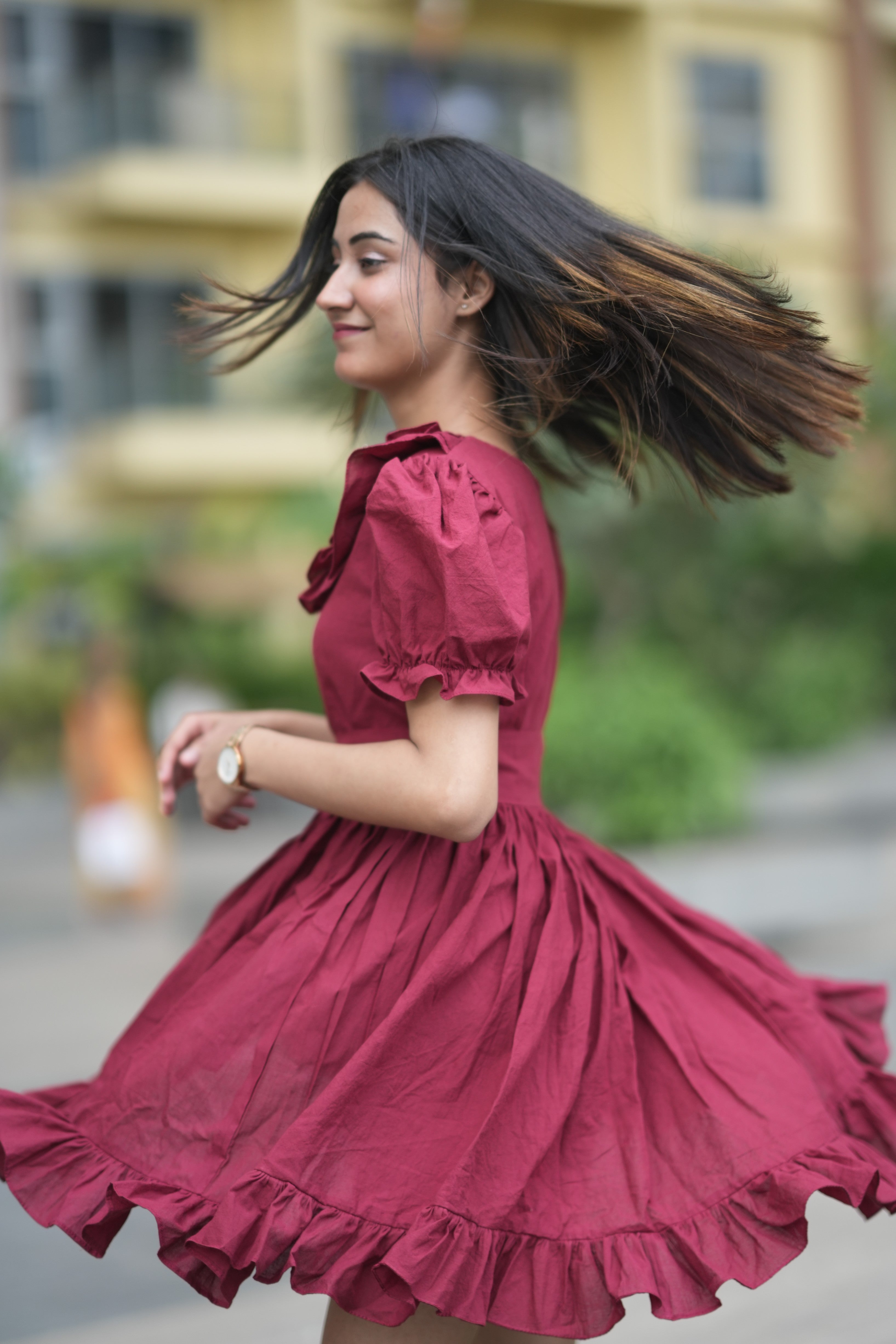 red ruffle dress, organic cotton mini dress, dress with puff sleeves, waist tie dress, fit and flare dress, vintage inspired clothing, sustainable mini dress, dress with built-in shorts and pockets, Swaruchii Veira, ruby red dress. A woman wearing The Ruby Ruffle Dress, a short ruby-red dress with puff sleeves, a ruffled neckline, and a matching fabric tie-belt, twirling outdoors with a joyful expression.
