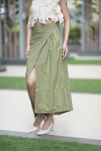Person wearing a green skirt with a white top in an outdoor setting. A model showcases 'The Cascade' co-ord set from Swaruchii, featuring a cream one-shoulder ruffled top and a high-waisted olive green Khadi wrap skirt with a high slit.