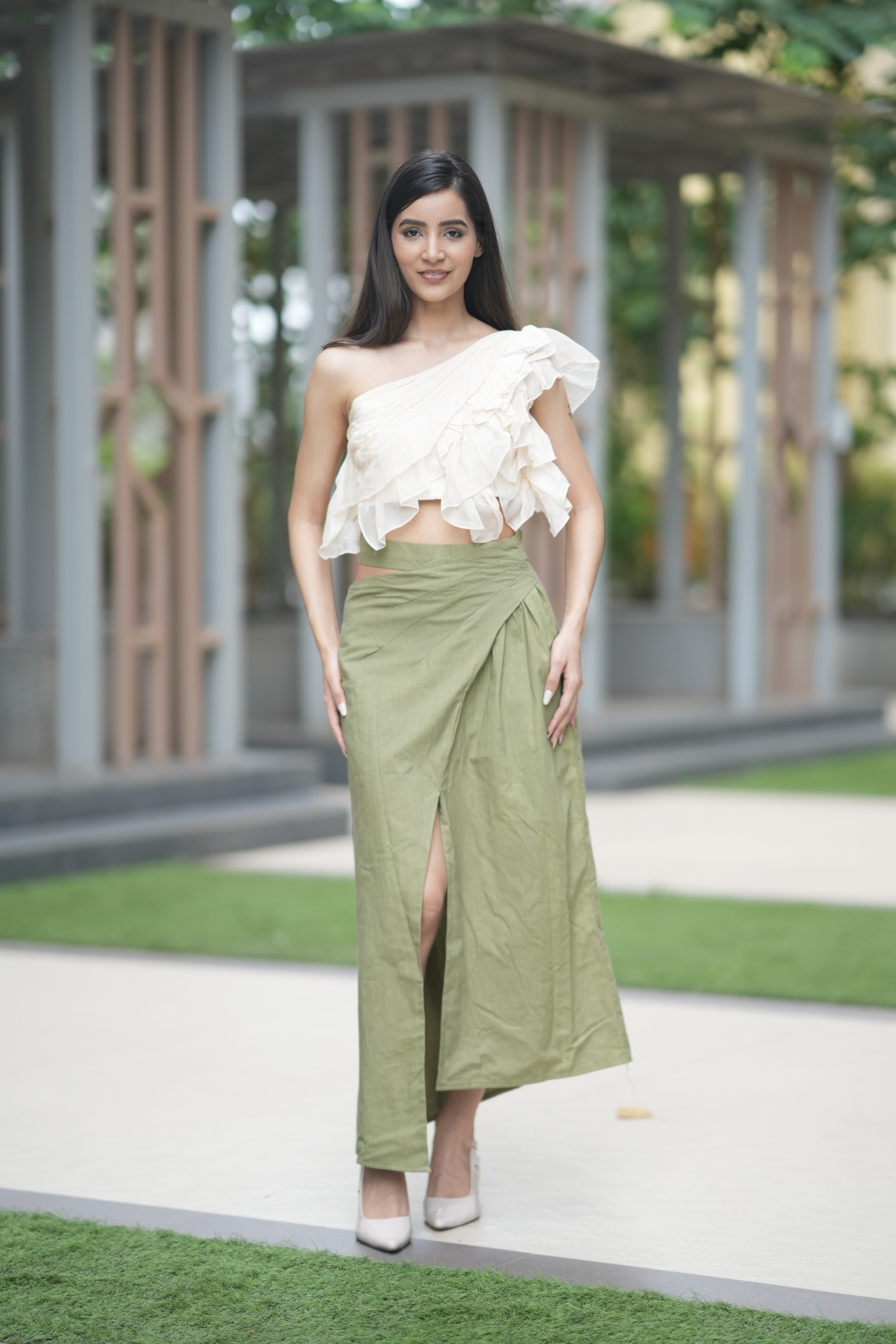Woman wearing a white one-shoulder top and green skirt standing outdoors. A model showcases 'The Cascade' co-ord set from Swaruchii, featuring a cream one-shoulder ruffled top and a high-waisted olive green Khadi wrap skirt with a high slit.