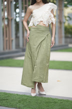 Woman wearing a white one-shoulder top and green skirt outdoors. A model showcases 'The Cascade' co-ord set from Swaruchii, featuring a cream one-shoulder ruffled top and a high-waisted olive green Khadi wrap skirt with a high slit.
