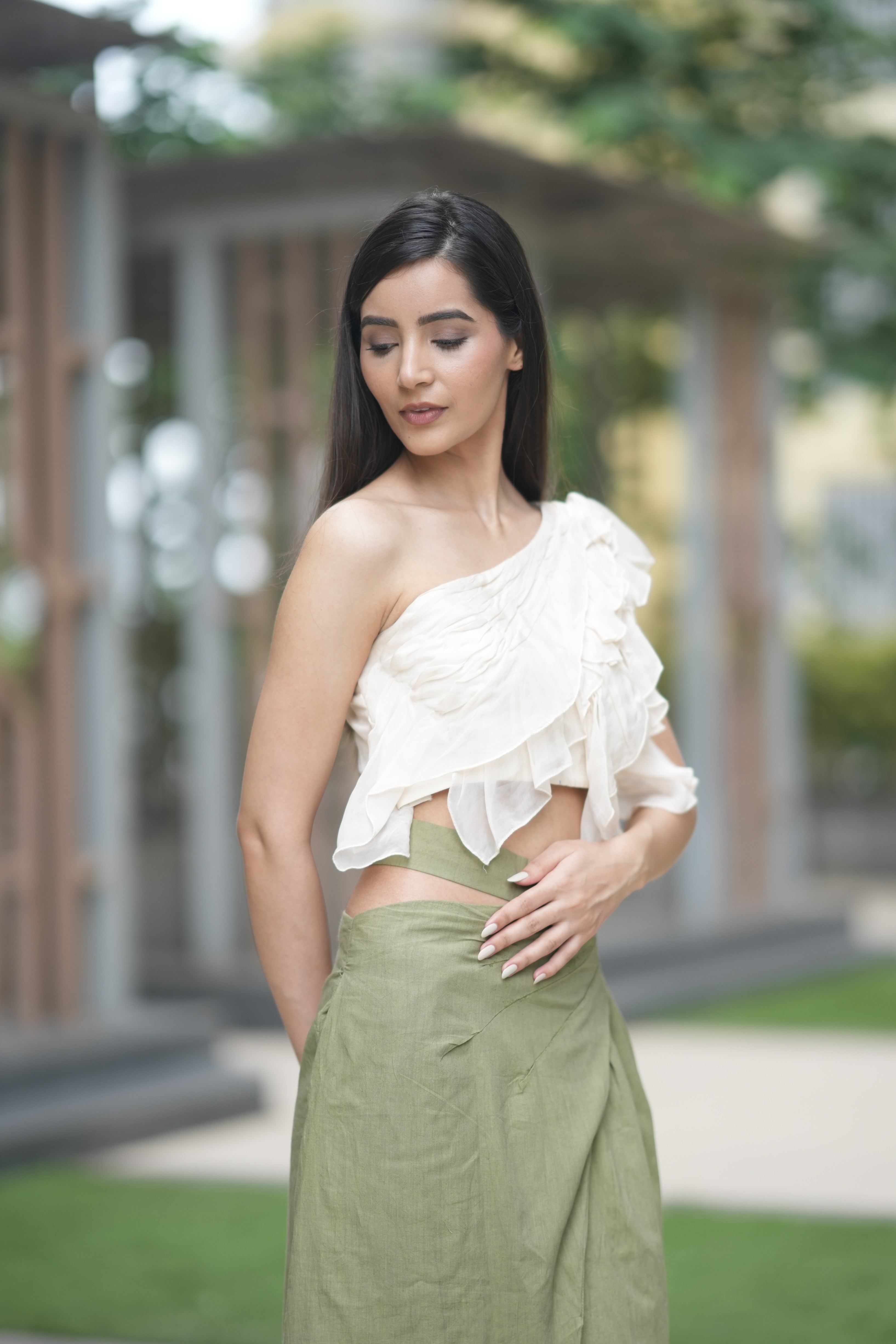 Woman wearing a white one-shoulder top and green skirt outdoors. A model showcases 'The Cascade' co-ord set from Swaruchii, featuring a cream one-shoulder ruffled top and a high-waisted olive green Khadi wrap skirt with a high slit.