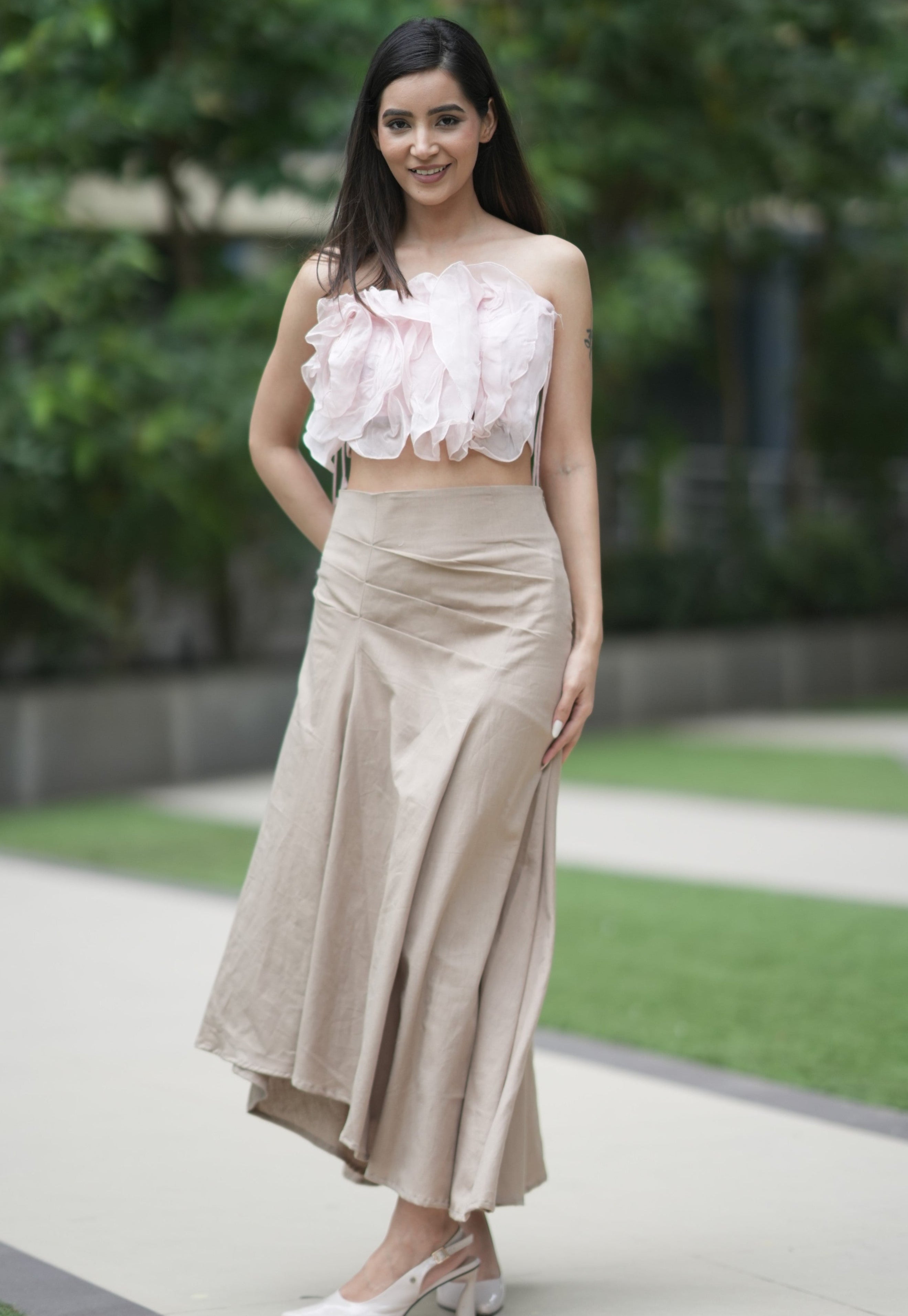 Woman wearing a floral top and beige skirt standing outdoors with greenery in the background. A woman models 'The Blush' co-ord set by Swaruchii, consisting of a pink ruffled strapless crop top and a beige asymmetrical Khadi midi skirt.