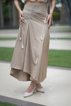 Person wearing a beige skirt with white tassels on a blurred outdoor background. A woman models 'The Blush' co-ord set by Swaruchii, consisting of a pink ruffled strapless crop top and a beige asymmetrical Khadi midi skirt.